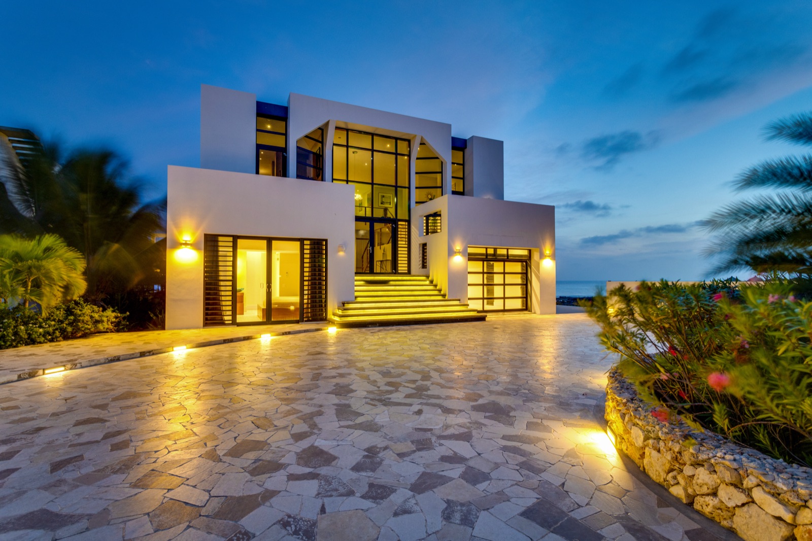 Modern luxury Bonaire villa exterior at twilight with an illuminated grand entrance, expansive glass windows, and a natural stone driveway overlooking the ocean.